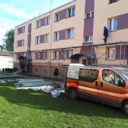 Pracownik maluje elewację trzypiętrowego budynku z rusztowania, widoczny pomarańczowy samochód dostawczy firmy i materiały budowlane na trawniku.