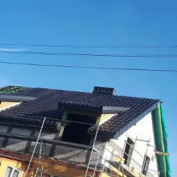 Dom w trakcie prac elewacyjnych z widocznym rusztowaniem, częściowo ocieploną i otynkowaną ścianą, oraz nowym dachem z kominem na tle błękitnego nieba.