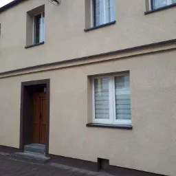 Beżowa elewacja domu jednorodzinnego z brązowymi akcentami wokół okien, drzwi i u podstawy, widoczna kostka brukowa na chodniku.