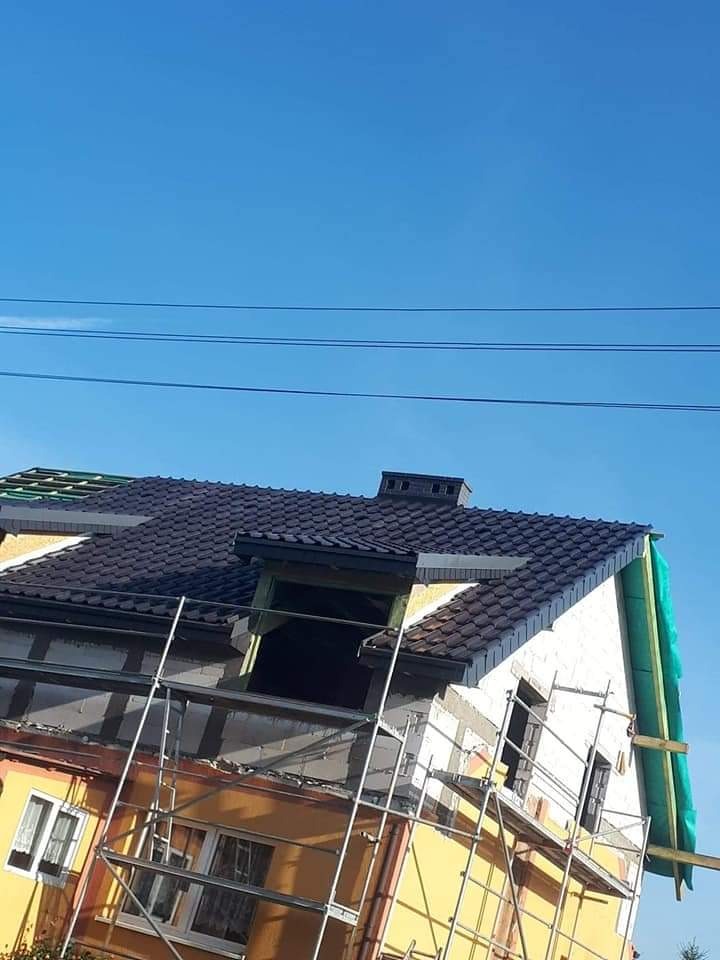 Dom w trakcie prac elewacyjnych z widocznym rusztowaniem, częściowo ocieploną i otynkowaną ścianą, oraz nowym dachem z kominem na tle błękitnego nieba.