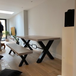 Madostal - Drewniany stół i ławka z czarnymi metalowymi nogami w kształcie litery X w nowoczesnym wnętrzu, widoczne miski dla zwierząt przy ścianie.