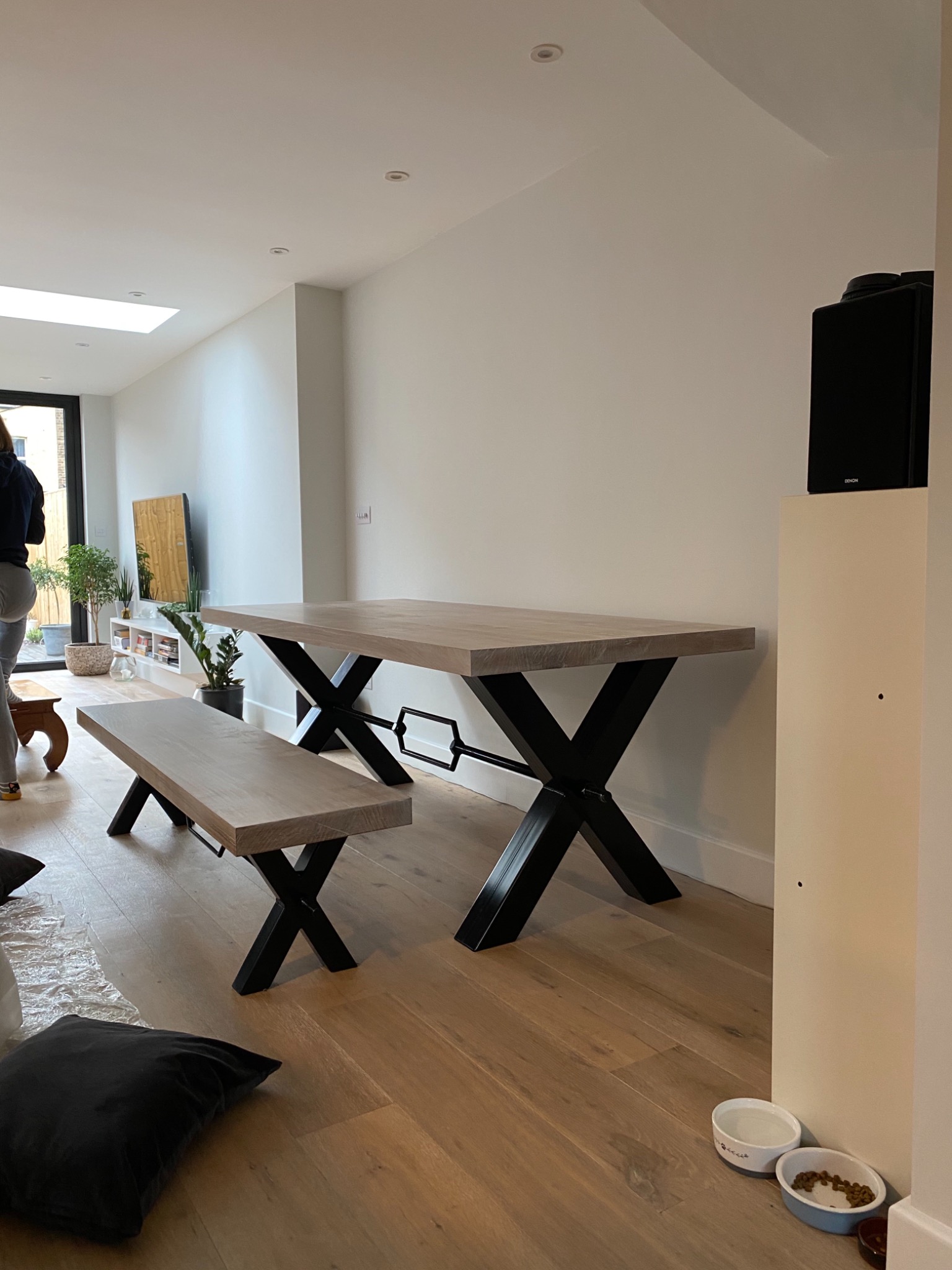 Drewniany stół i ławka z czarnymi metalowymi nogami w kształcie litery X w nowoczesnym wnętrzu, widoczne miski dla zwierząt przy ścianie.