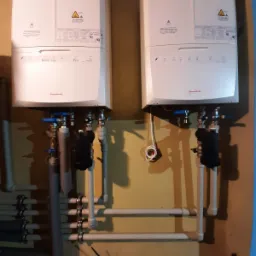 Dwa identyczne, białe kotły grzewcze marki Immergas zamontowane na żółtej ścianie, z widocznymi rurami i osprzętem hydraulicznym, oraz czerwonym laserowym miernikiem odległości u dołu.