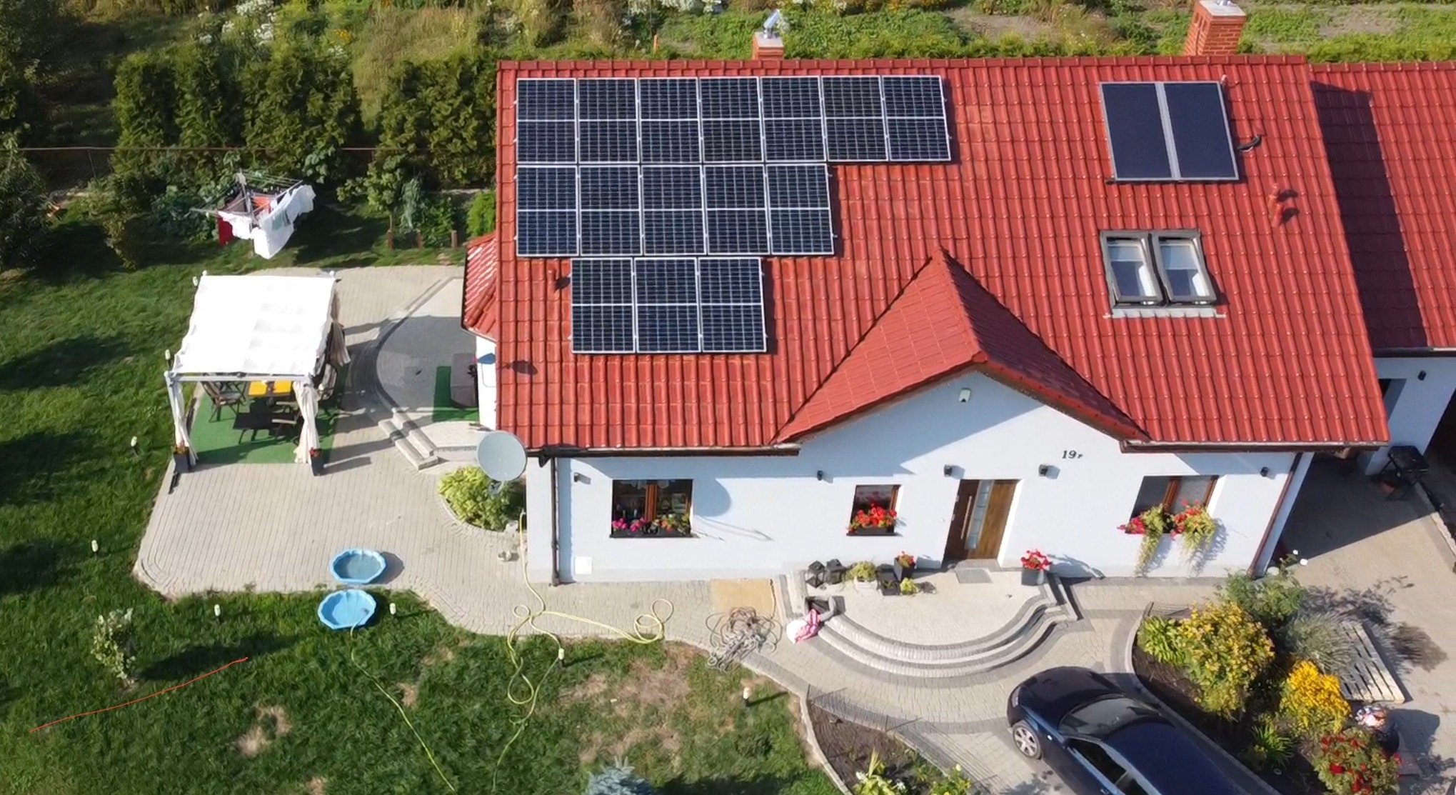 Widok z lotu ptaka na dom jednorodzinny z czerwoną dachówką, na której zamontowano panele słoneczne; obok domu widoczny ogród z altaną, dwa małe baseny oraz zaparkowany samochód.