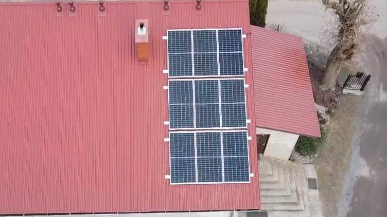 Widok z góry na panele słoneczne zamontowane na czerwonym blaszanym dachu budynku mieszkalnego z kominem.