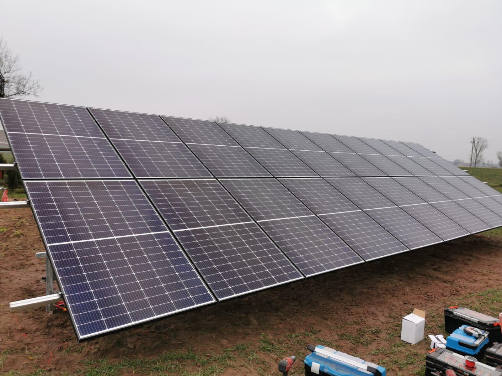 Instalacja fotowoltaiczna: rzędy paneli słonecznych zamontowane na ziemi, widoczne narzędzia i opakowania w trakcie montażu, pochmurne niebo w tle.