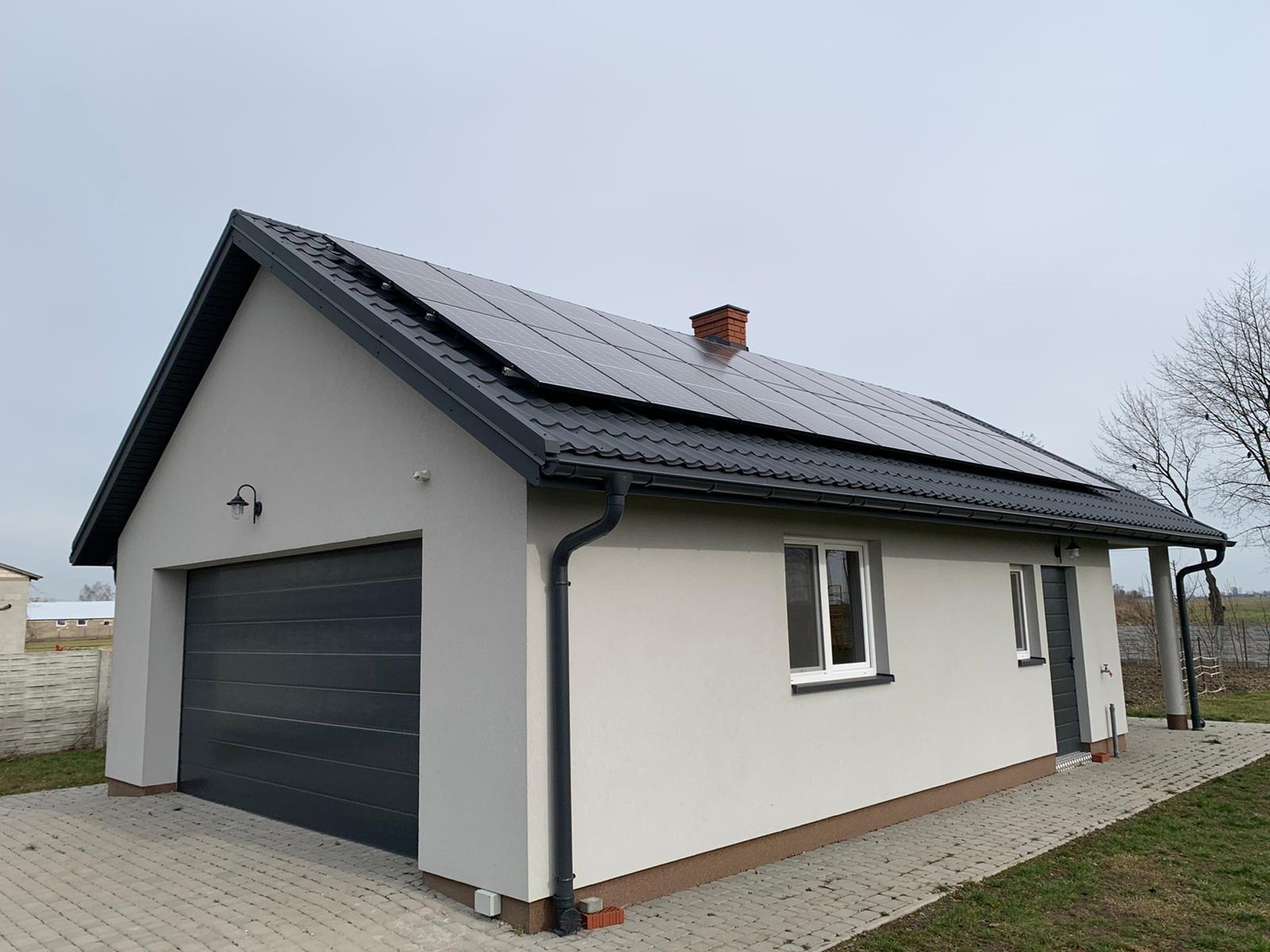 Nowoczesny, parterowy budynek z szarą bramą garażową i panelami słonecznymi na dachu pokrytym blachodachówką, widoczny komin z cegły i rynny w kolorze dachu.