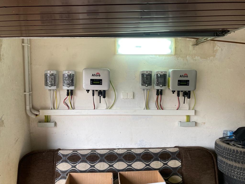 Rząd falowników marki Afore zamontowanych na białej listwie wewnątrz budynku z widocznymi kablami i instalacją elektryczną, poniżej widoczny fragment sofy i opon.