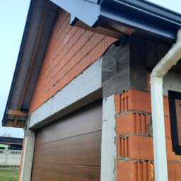 RYNNY PROSTOKĄTNE ZBIGNIEW CZUTRO - Niedokończona elewacja budynku z brązową bramą garażową, widoczna orynnowana część dachu z szarą blachodachówką, biała rura spustowa i surowy mur z czerwonej cegły dziurawki.