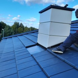 RYNNY PROSTOKĄTNE ZBIGNIEW CZUTRO - Montaż obudowy komina na dachu z ciemnej dachówki ceramicznej, widoczny fragment pracownika w szarym ubraniu i fragment narzędzia, błękitne niebo w tle.