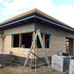 RYNNY PROSTOKĄTNE ZBIGNIEW CZUTRO - Budynek w trakcie budowy z zamontowanymi, grafitowymi rynnami. Widoczna drabina oparta o ścianę, palety z dachówką Braas i niezadaszona konstrukcja dachu.