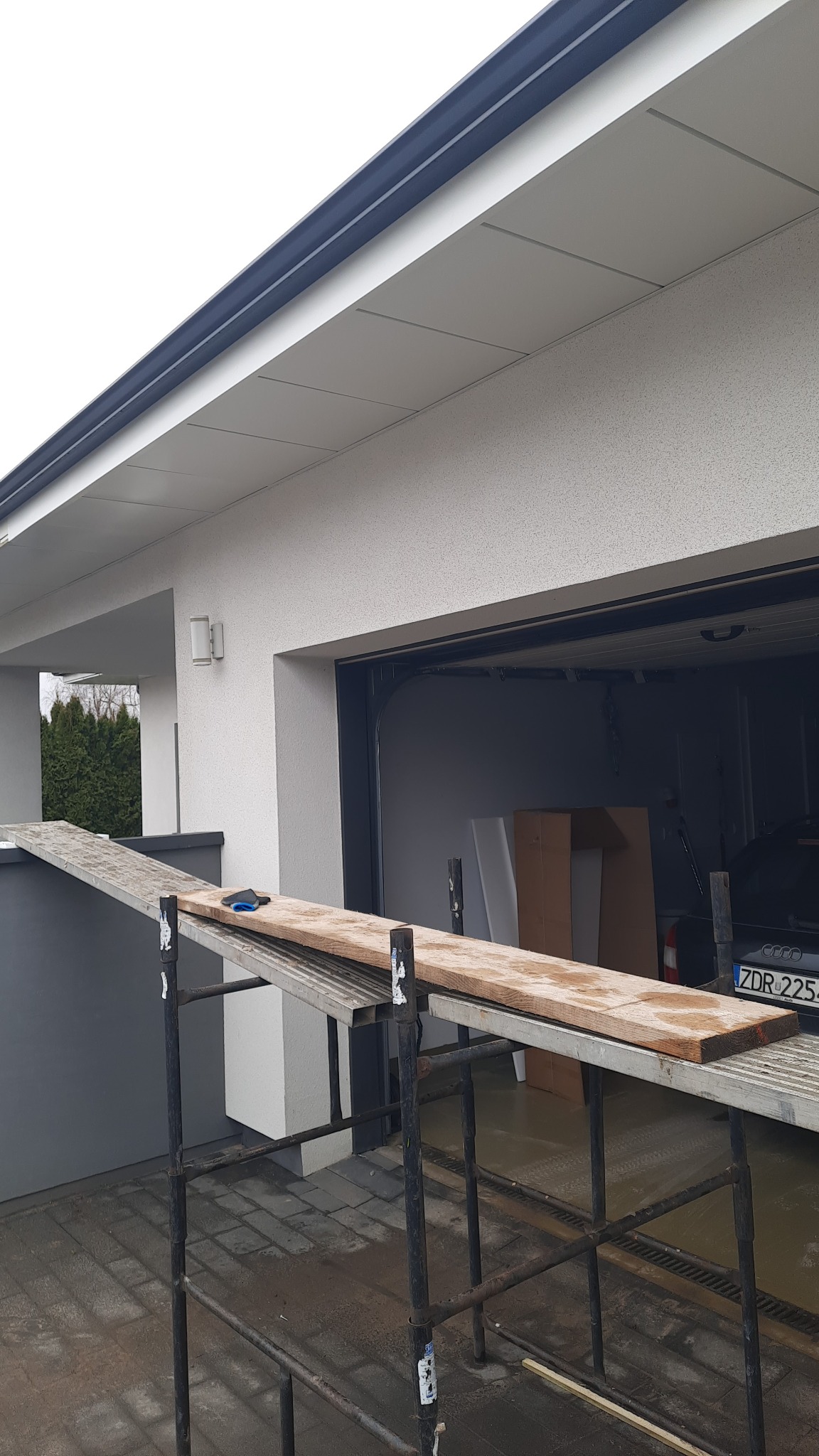 Szara rynna zamontowana przy jasnej elewacji domu w Złocieńcu, widoczna z poziomu rusztowania z deską i rękawicami roboczymi.