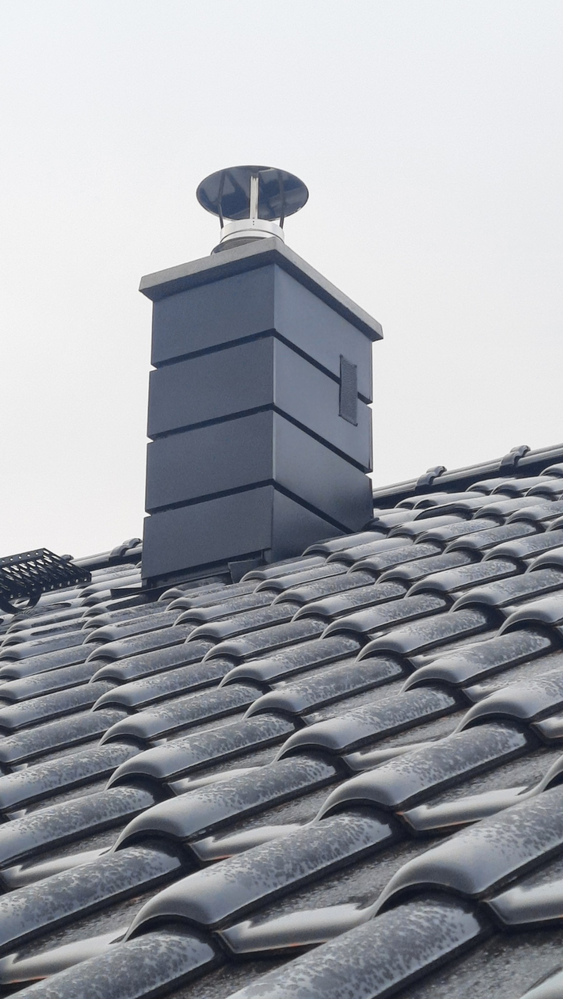 Czarny, metalowy komin z daszkiem na błyszczącym, czarnym dachu z dachówki ceramicznej, widziany pod kątem w pochmurny dzień.