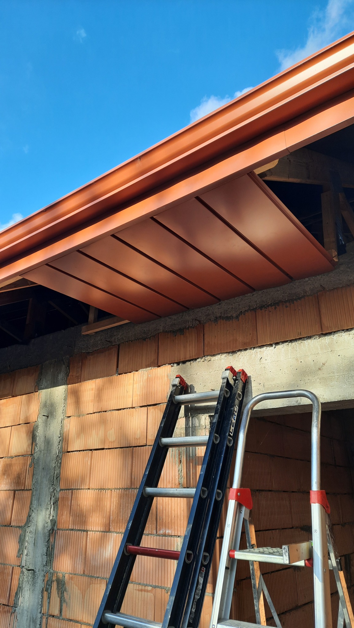Montaż okapu dachowego z metalowymi panelami w kolorze miedzianym, widoczne dwie drabiny opierające się o ścianę budynku z pustaków ceramicznych, błękitne niebo w tle.