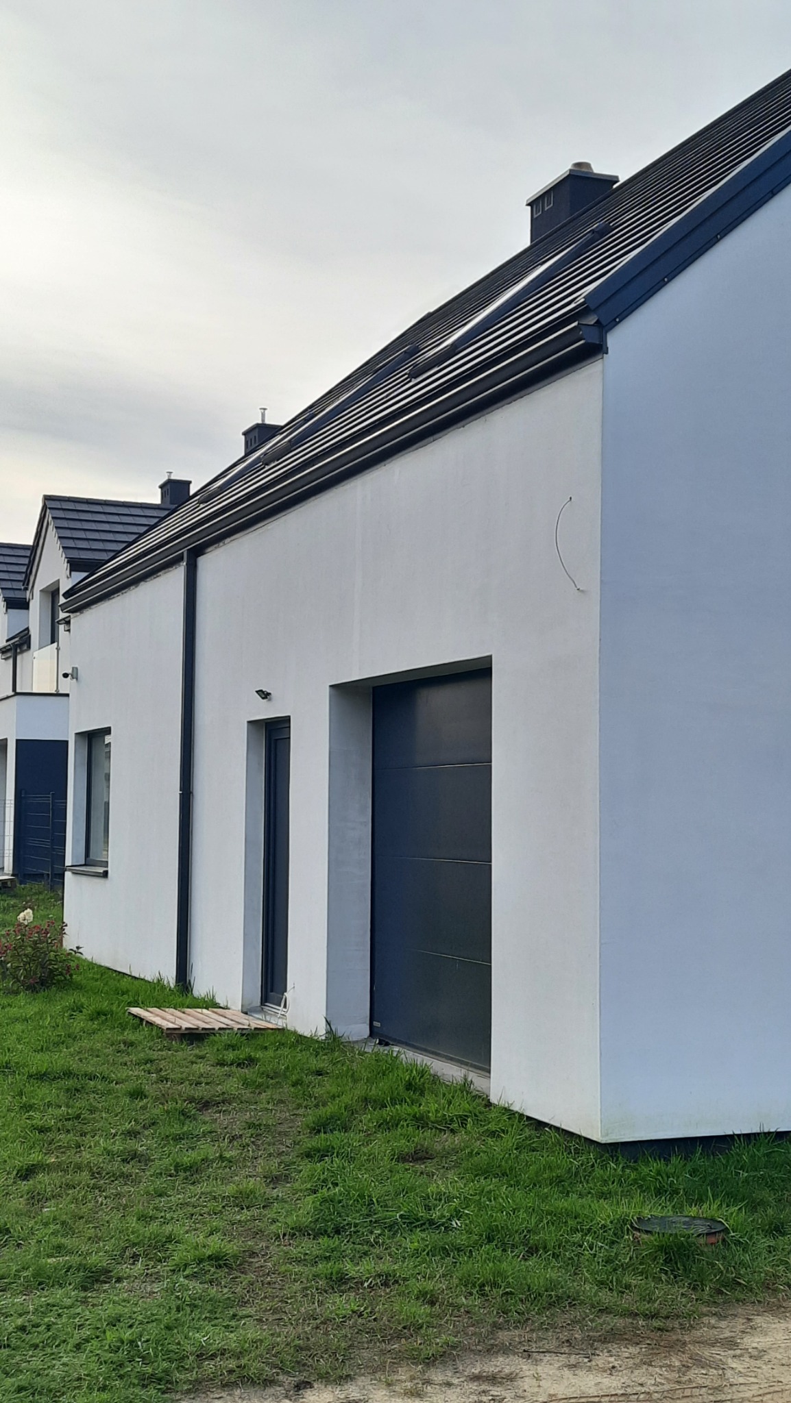 Nowoczesny dom jednorodzinny z białymi ścianami, grafitową stolarką okienną i rynnami w kolorze dachu, widok z poziomu trawnika.