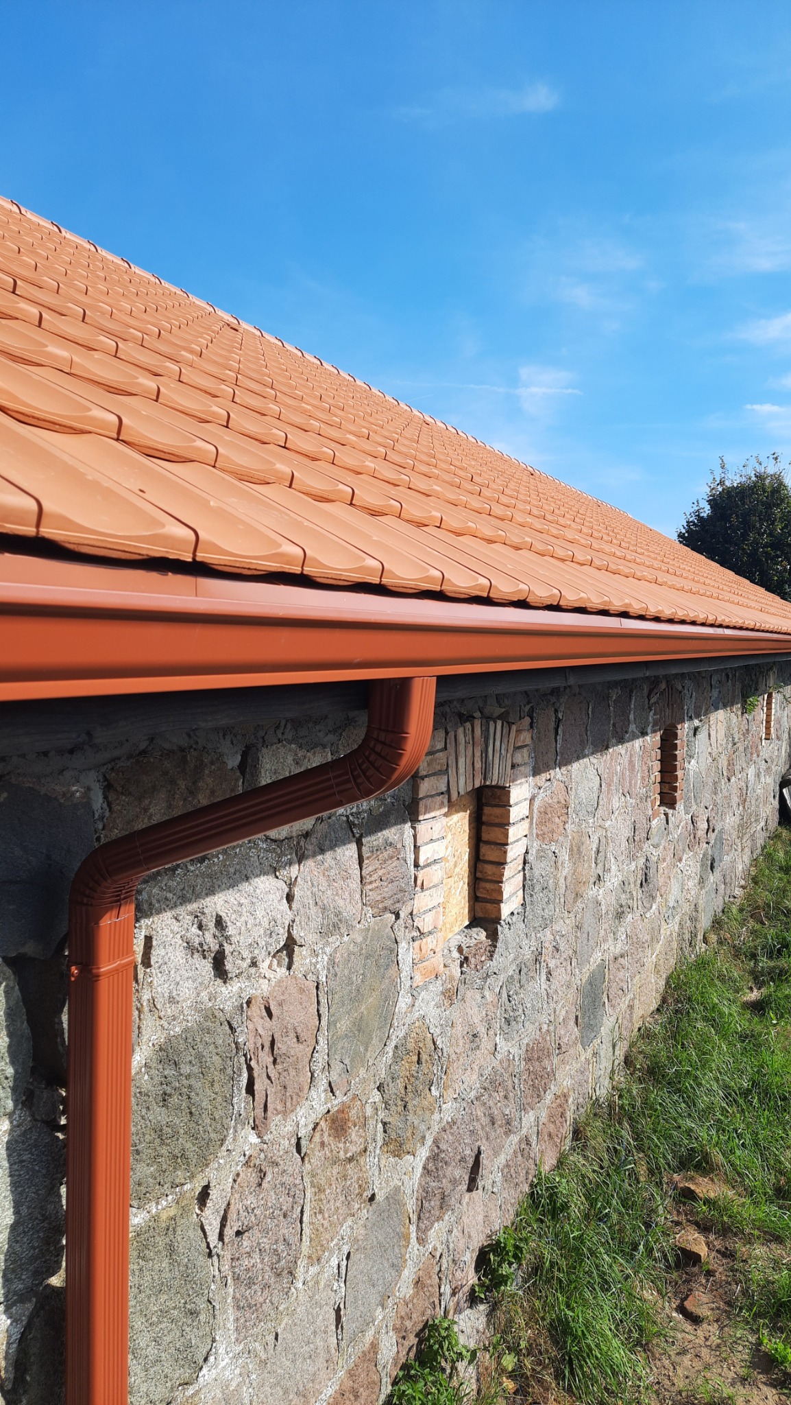 Pomarańczowa rynna spustowa zamontowana na kamiennej ścianie budynku z ceglanymi wstawkami, widoczny fragment dachu z dachówki w tym samym kolorze, błękitne niebo w tle.