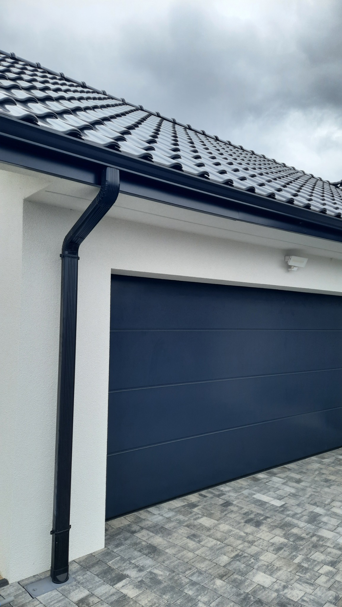 Nowoczesny dom z czarną, metalową rynną i ciemnoszarą bramą garażową na tle pochmurnego nieba.