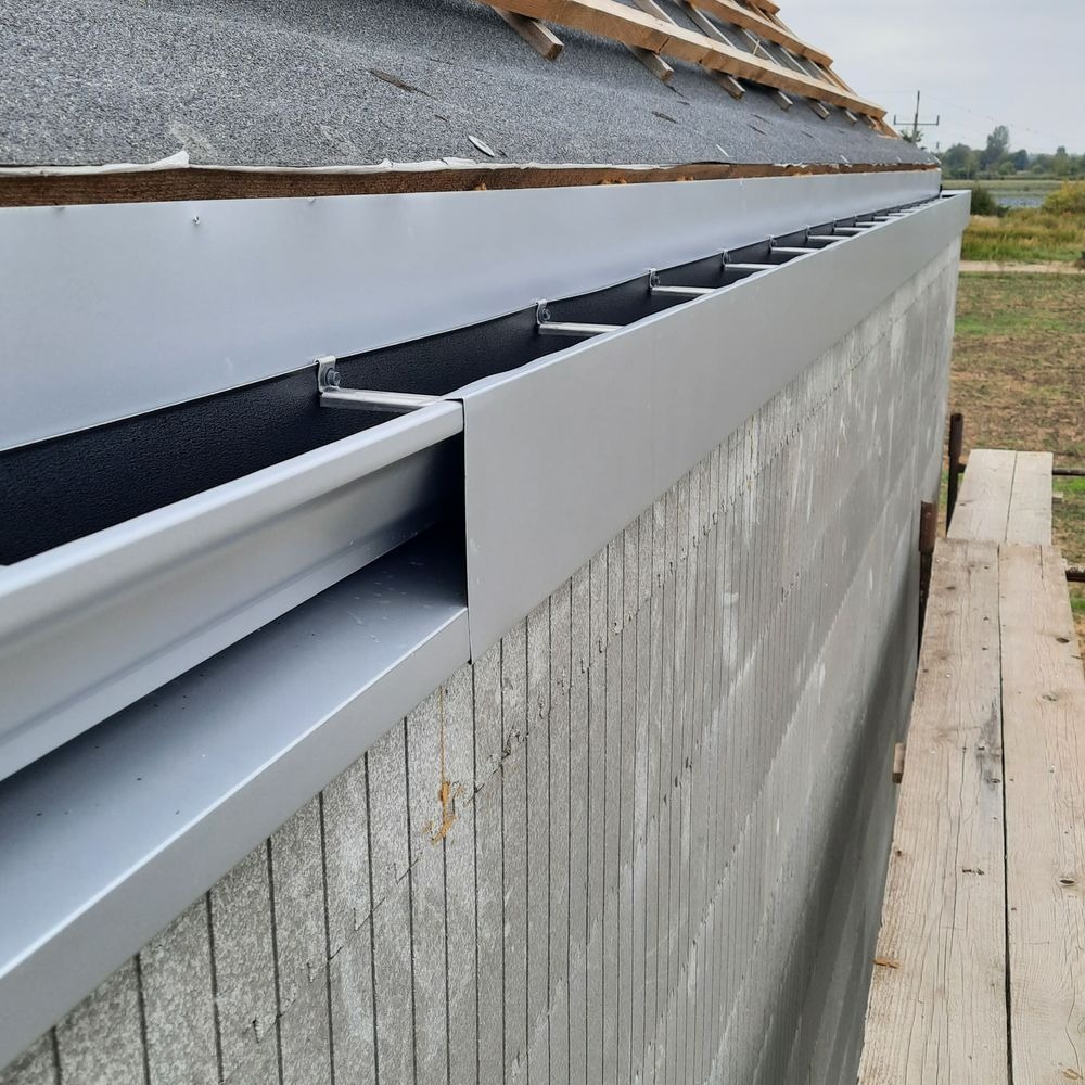 Nowe, srebrne orynnowanie zamontowane na szarym, surowym budynku z widocznym fragmentem dachu pokrytego papą i elementami drewnianej konstrukcji.