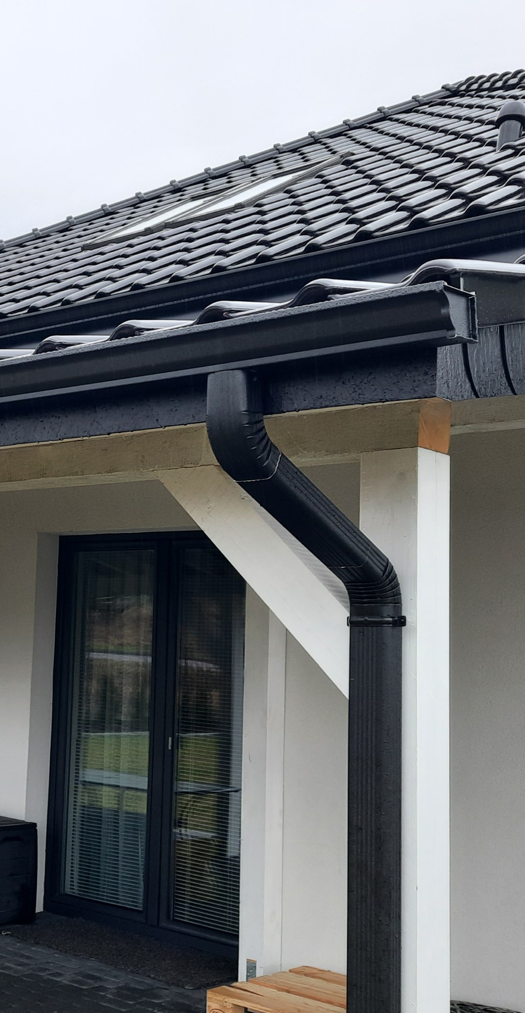 Czarna, metalowa rynna spustowa zamontowana przy nowoczesnym domu z ciemną dachówką i oknami. Widoczny fragment elewacji z drewnianym elementem konstrukcyjnym.