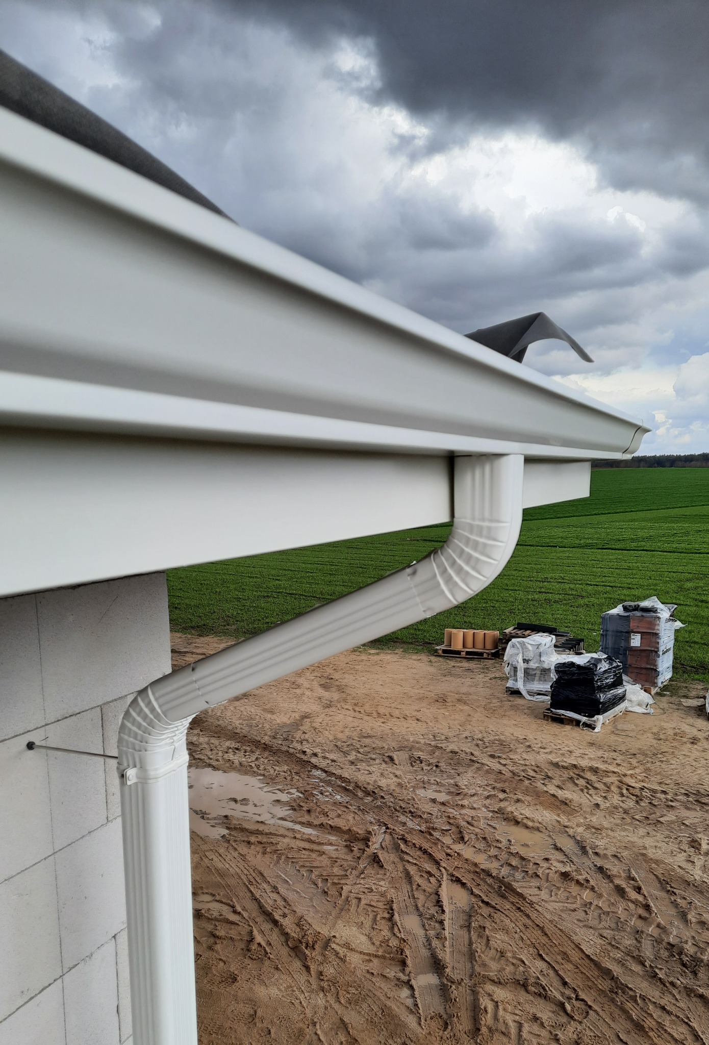 Nowo zamontowany, biały system odprowadzania wody deszczowej na tle zielonego pola i pochmurnego nieba. Widoczne elementy mocujące do ściany z bloczków betowych.