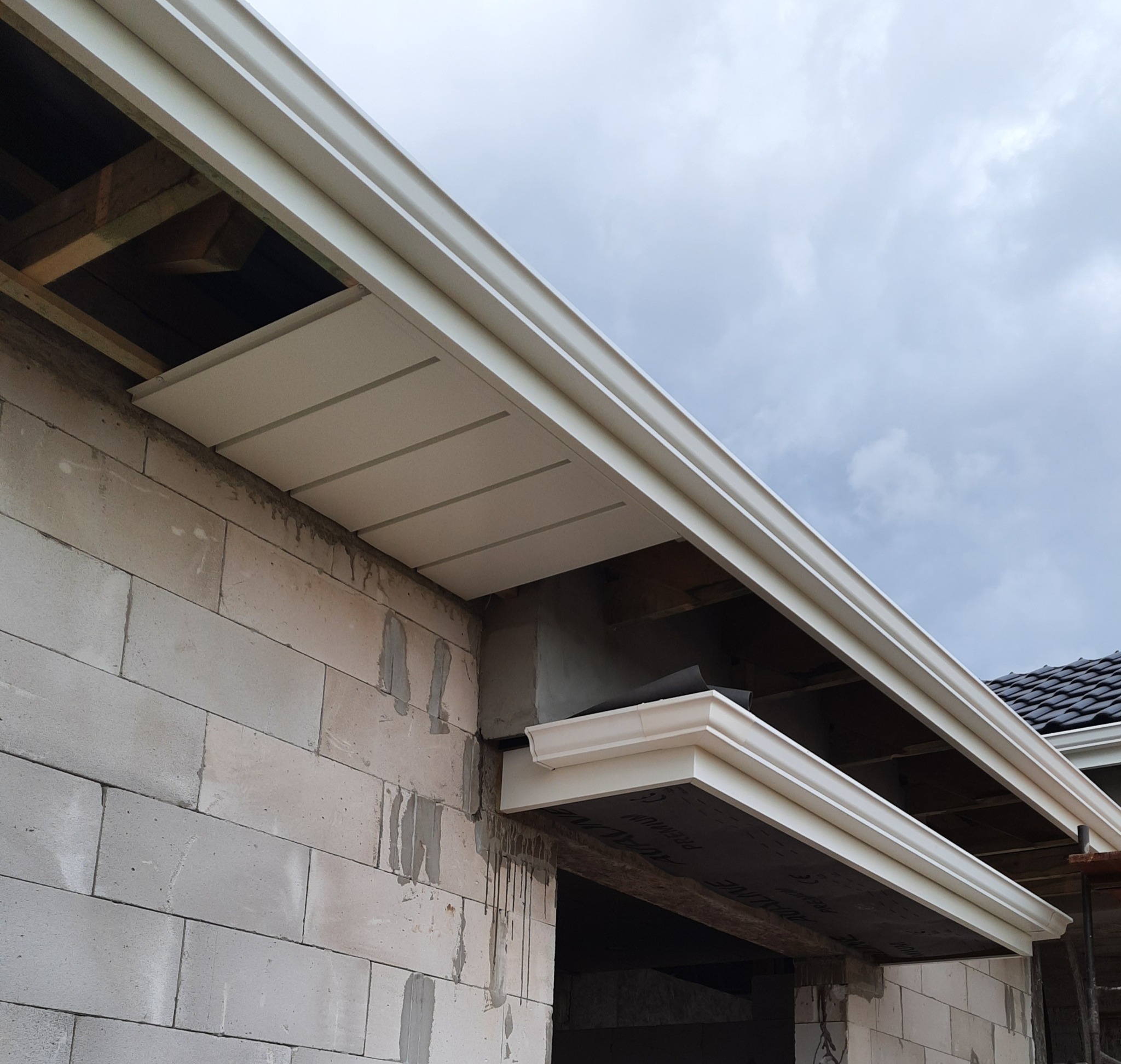 Białe rynny zamontowane na budynku z białych pustaków, widok od dołu na fragment dachu i pochmurne niebo.
