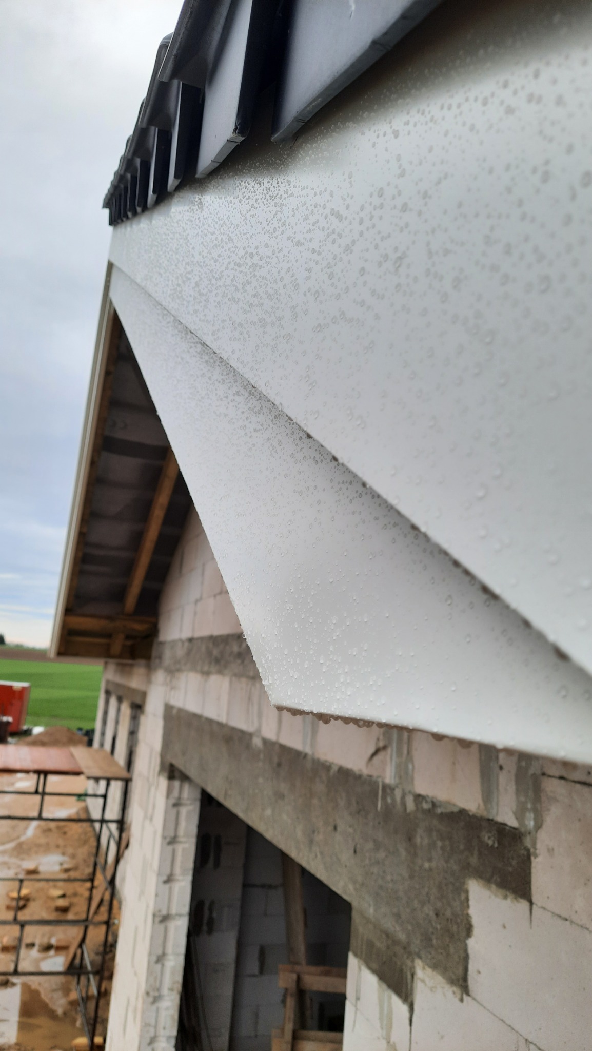 Biała rynna zamontowana na surowym budynku w trakcie budowy, pokryta kroplami deszczu, widok z dołu.
