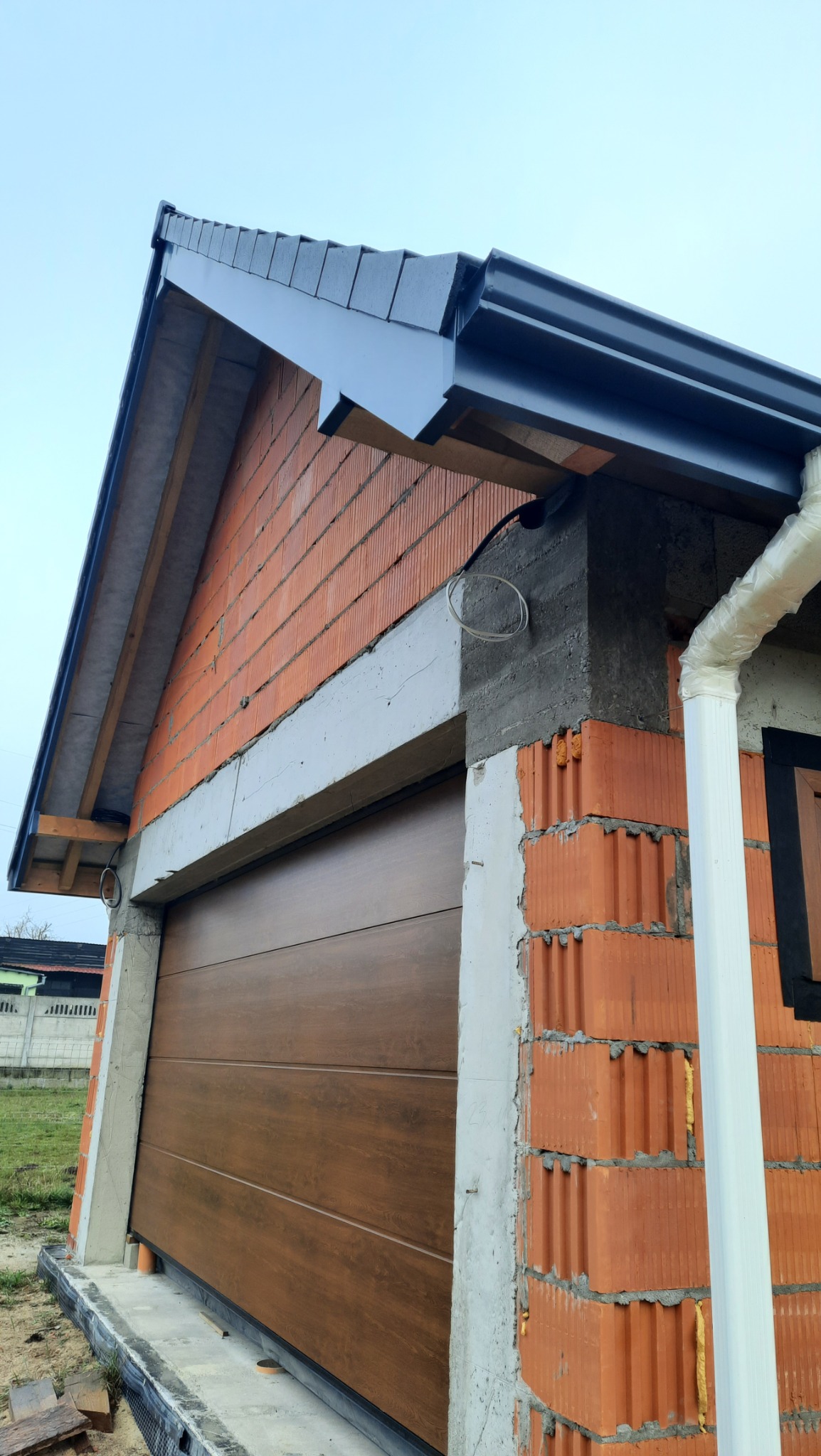 Niedokończona elewacja budynku z brązową bramą garażową, widoczna orynnowana część dachu z szarą blachodachówką, biała rura spustowa i surowy mur z czerwonej cegły dziurawki.