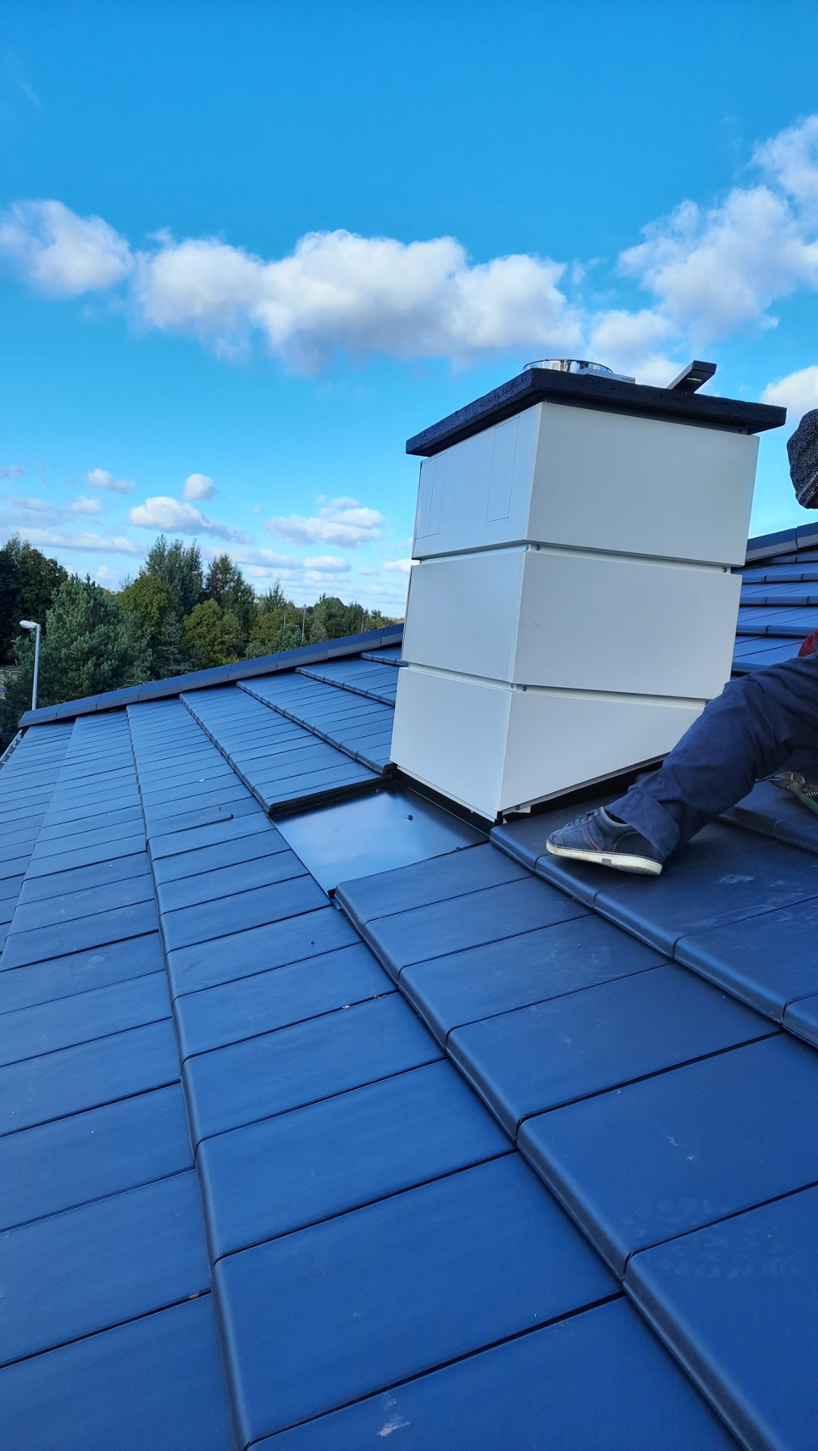 Montaż obudowy komina na dachu z ciemnej dachówki ceramicznej, widoczny fragment pracownika w szarym ubraniu i fragment narzędzia, błękitne niebo w tle.
