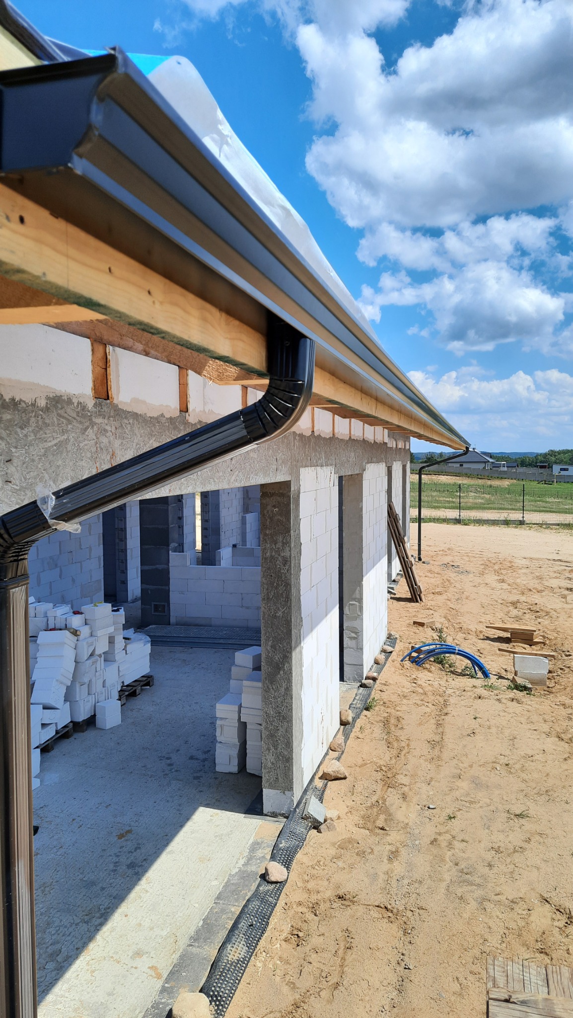 Czarna, metalowa rynna zamontowana na drewnianej konstrukcji dachu domu w trakcie budowy; widoczne białe bloczki ścian i jasne, błękitne niebo z chmurami.