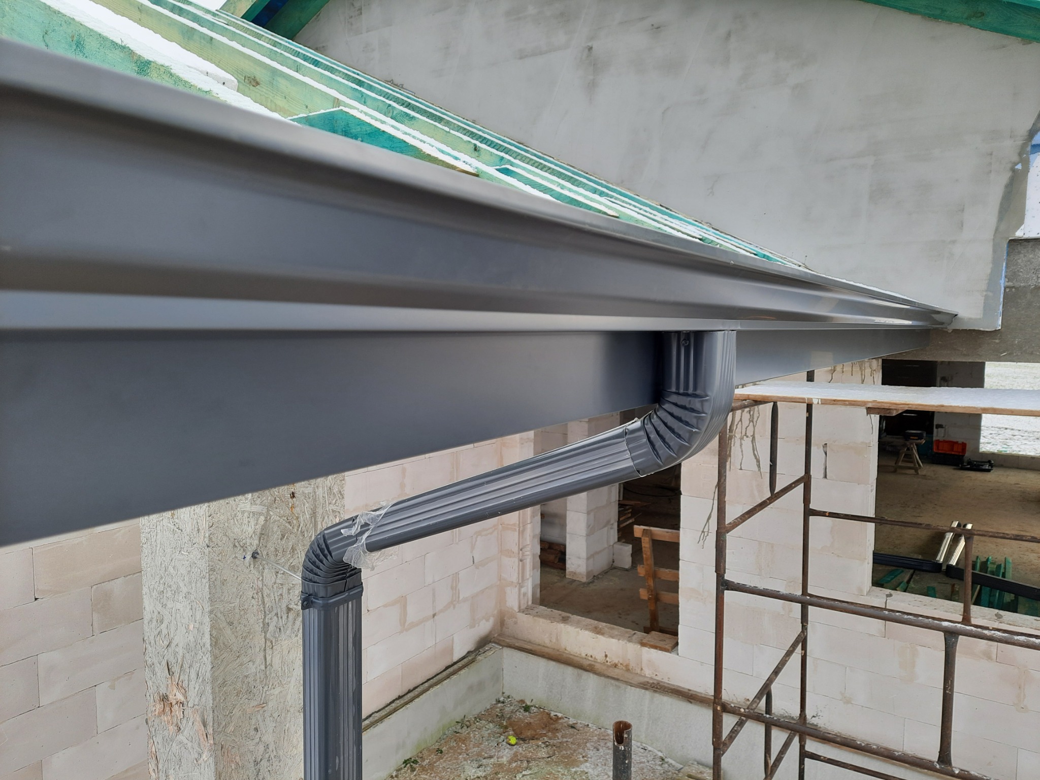 Szara rynna zamontowana na surowej elewacji budynku w trakcie budowy, widoczne elementy więźby dachowej i rusztowanie.