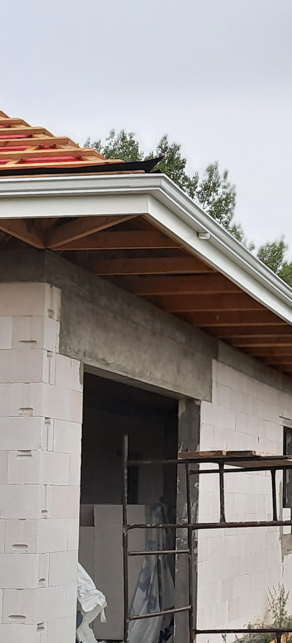 Widok z dołu na białą rynnę zamontowaną na nowym budynku z białych bloczków, widoczne krokwie dachu i rusztowanie.