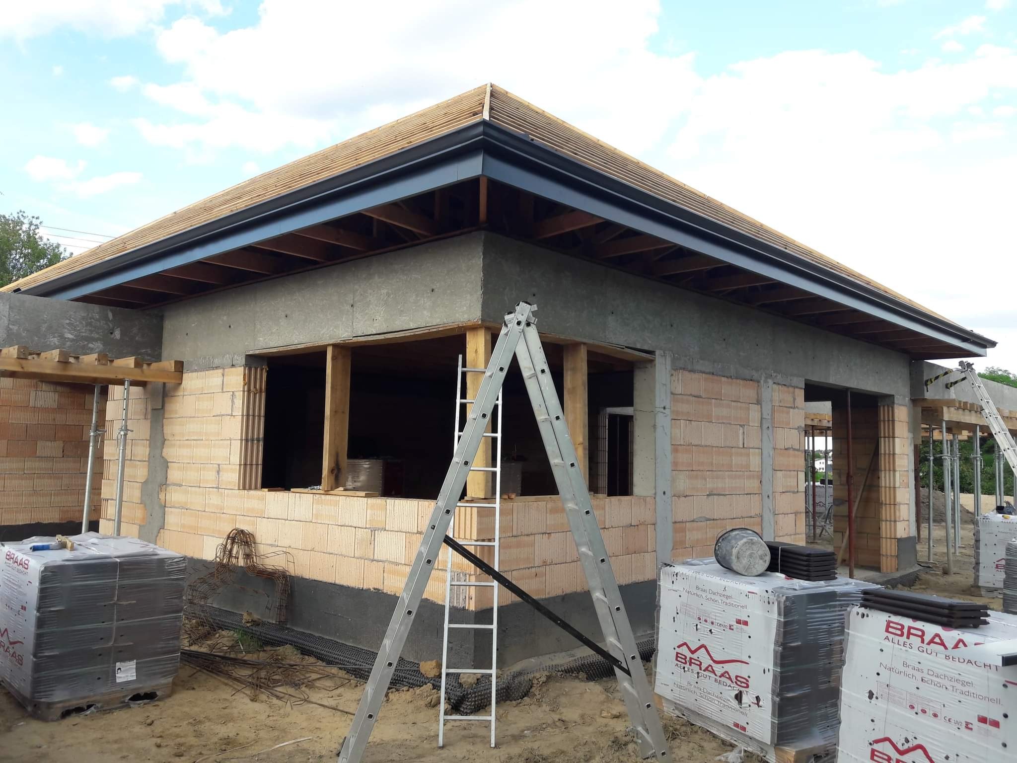 Budynek w trakcie budowy z zamontowanymi, grafitowymi rynnami. Widoczna drabina oparta o ścianę, palety z dachówką Braas i niezadaszona konstrukcja dachu.