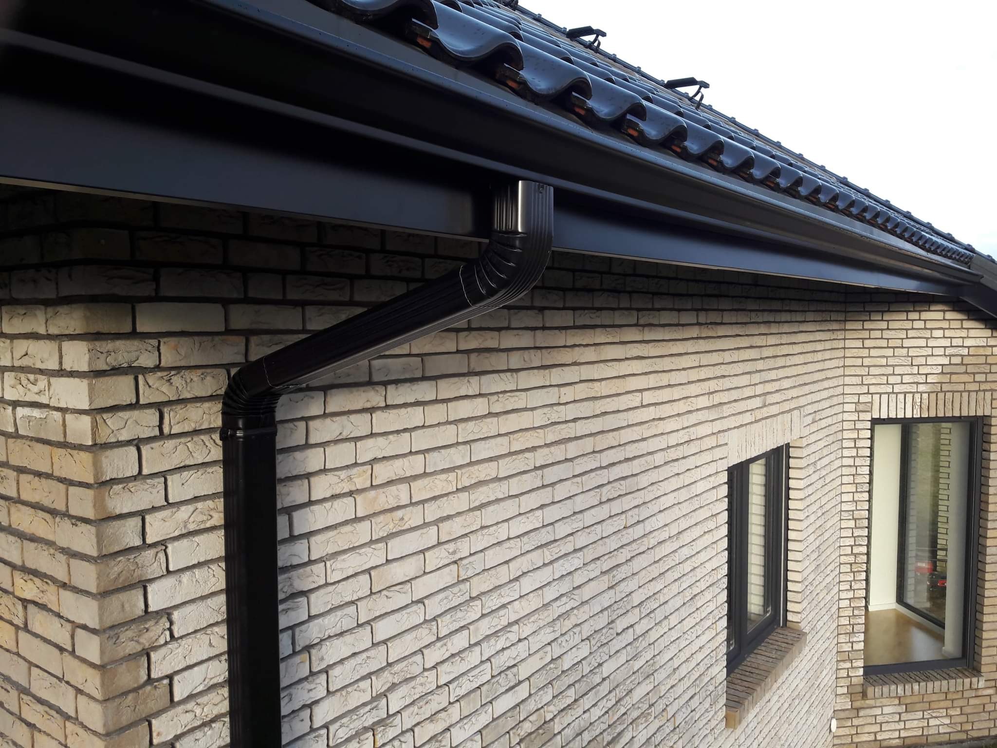 Zamontowany system rynnowy w kolorze czarnym na jasnej elewacji z cegły klinkierowej, widok z dołu na dach z czarną dachówką ceramiczną.