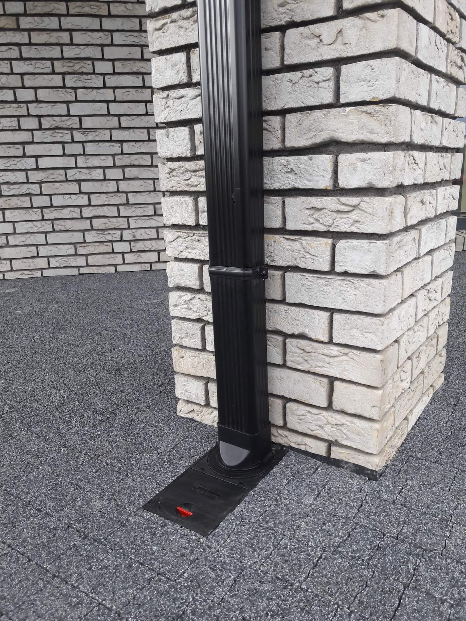 Czarna rura spustowa przy elewacji z jasnej cegły klinkierowej, z widocznym odpływem na szarym podłożu z kostki brukowej.