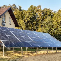 Instalacja fotowoltaiczna na metalowych podporach przed domem jednorodzinnym z ogrodem, panele odbijają błękitne niebo.