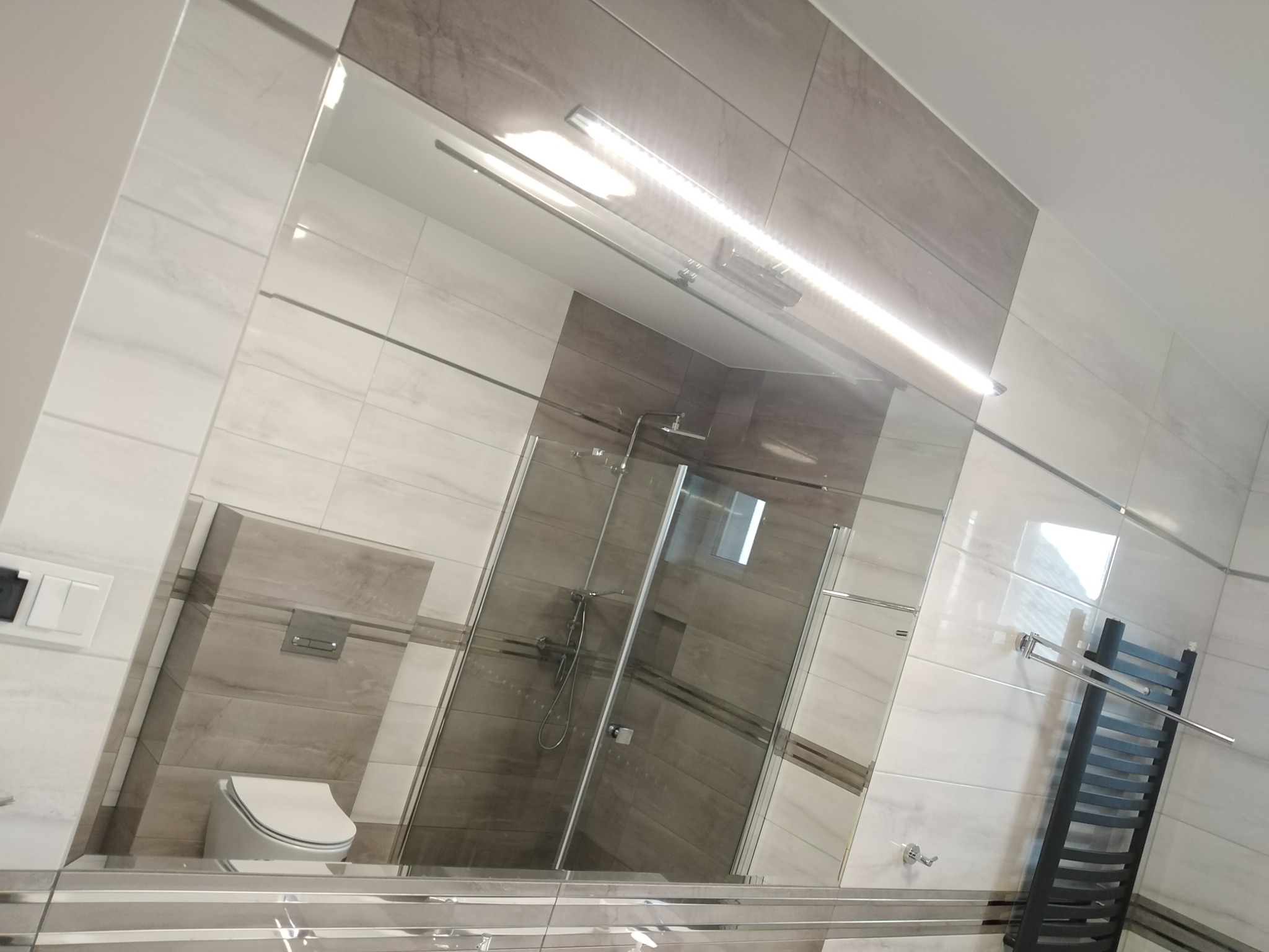 Nowoczesna łazienka z dużym lustrem, szarymi i białymi płytkami imitującymi marmur, kabiną prysznicową ze szklanymi drzwiami oraz czarnym grzejnikiem drabinkowym.