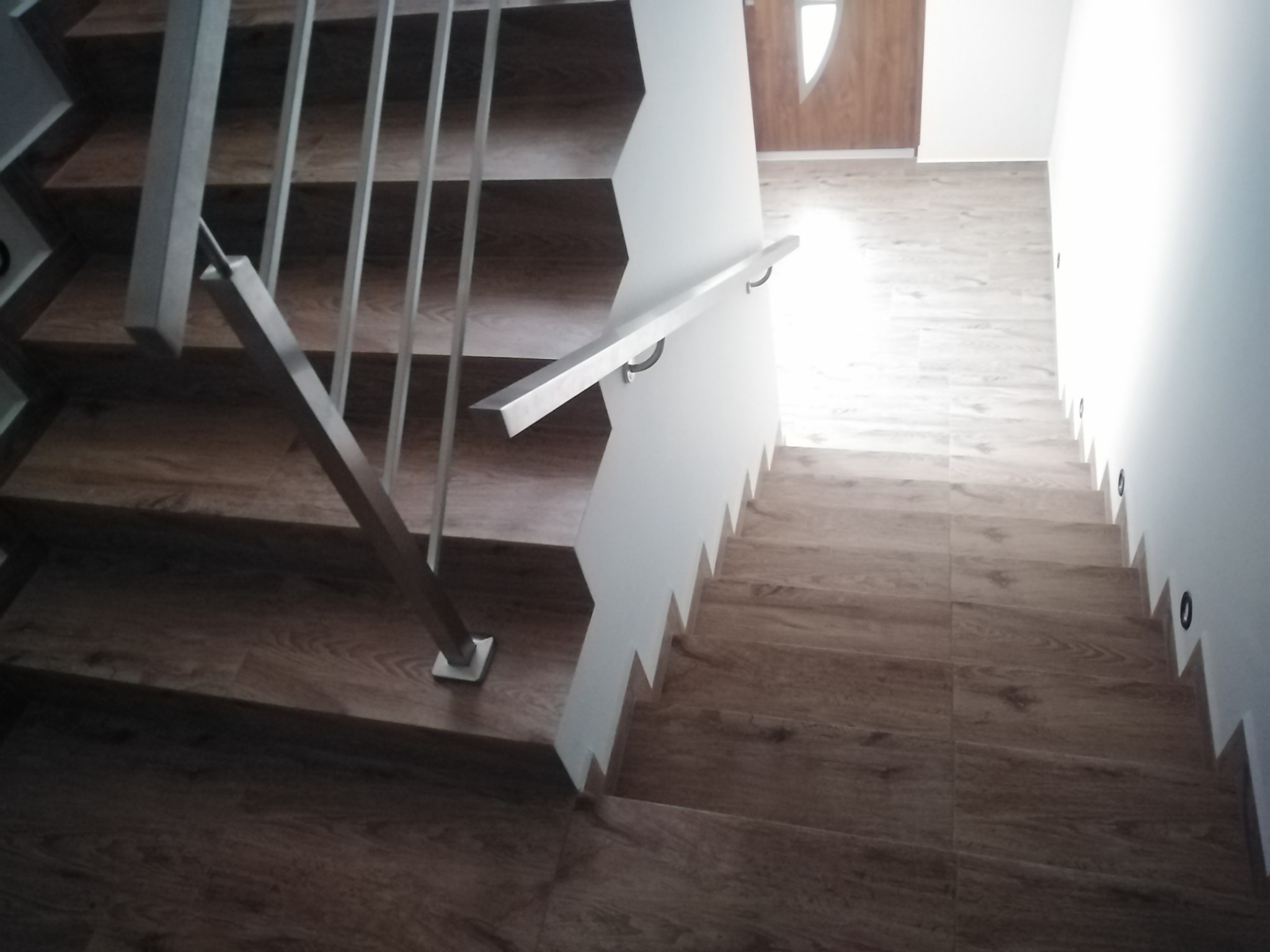 Schody wyłożone płytkami imitującymi drewno, z metalową balustradą i białymi ścianami, widok z góry, oświetlenie punktowe wzdłuż ściany.