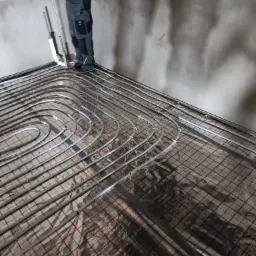 Instalacja ogrzewania podłogowego z rur na folii izolacyjnej z czerwoną siatką, widoczny pracownik z narzędziami w szarych spodniach roboczych, surowy stan ścian w tle.