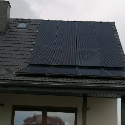 Panele słoneczne zamontowane na dachu domu z ciemnej dachówki, widoczne okno dachowe i komin. Dolne panele częściowo odbijają otoczenie.