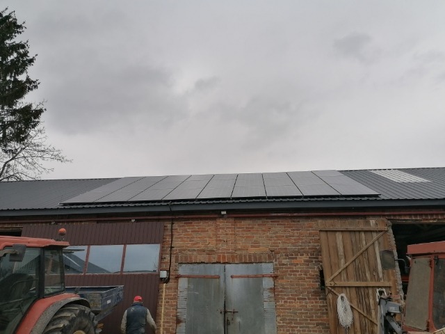 Panele słoneczne zamontowane na dachu budynku gospodarczego z czerwonej cegły, widoczne traktory i pochmurne niebo.