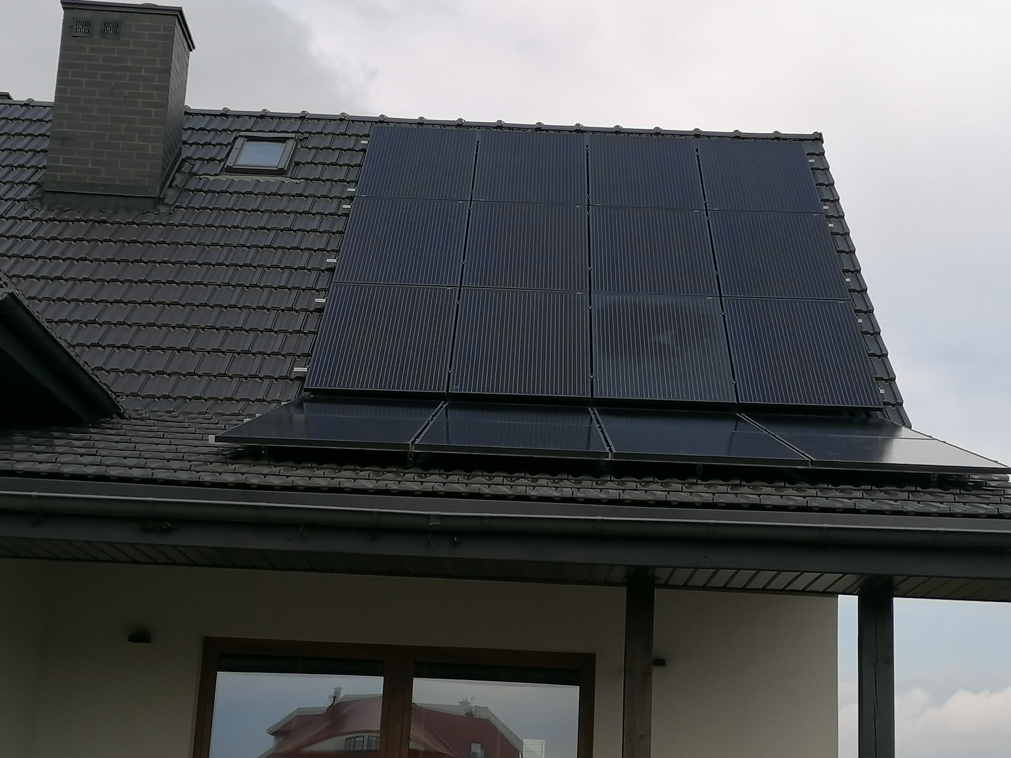 Panele słoneczne zamontowane na dachu domu z ciemnej dachówki, widoczne okno dachowe i komin. Dolne panele częściowo odbijają otoczenie.