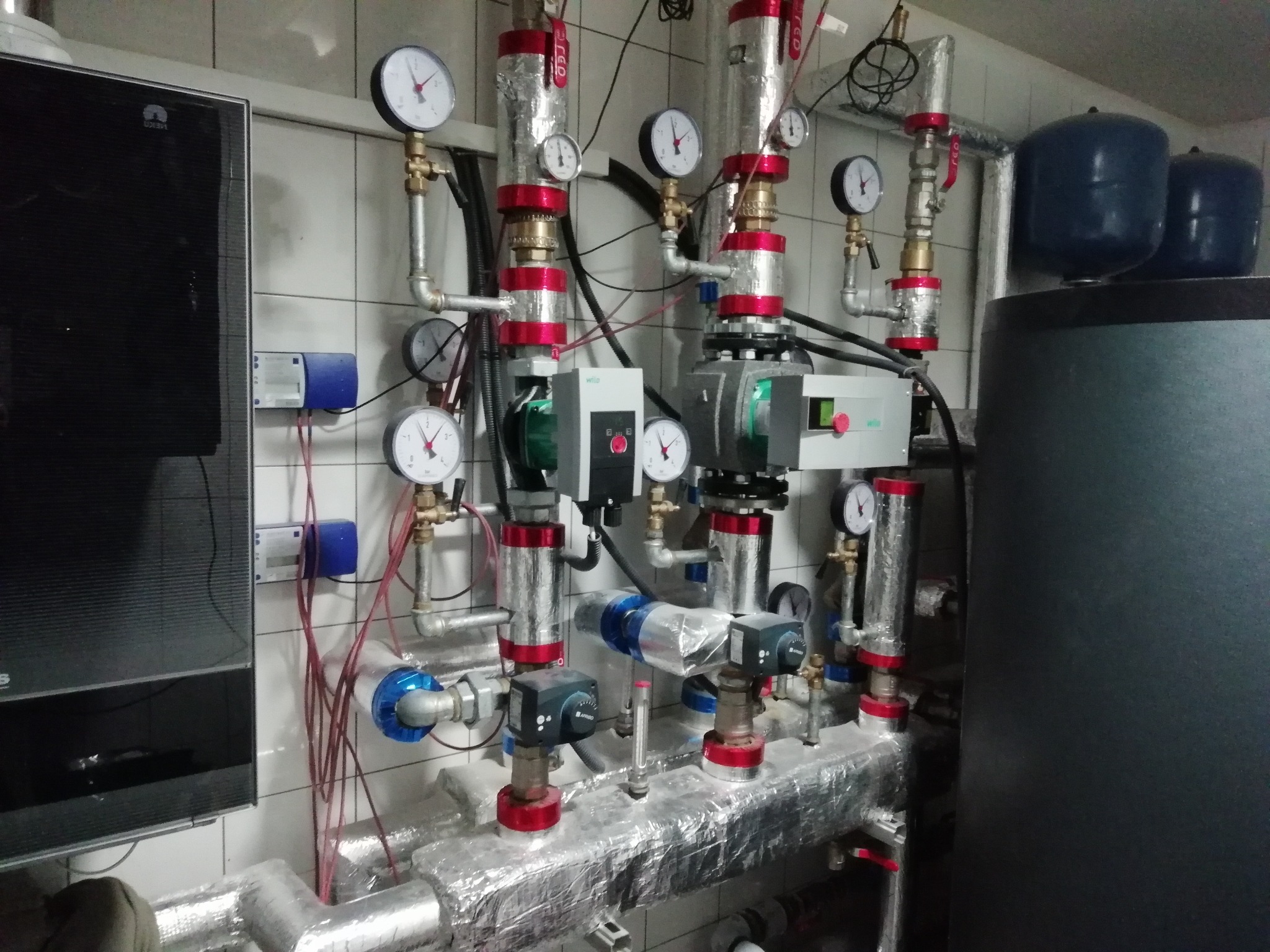 Złożona instalacja grzewcza z rurami owiniętymi folią izolacyjną, manometry, pompy obiegowe z regulacją, zbiorniki wyrównawcze, zamontowana na tle białych płytek ceramicznych.