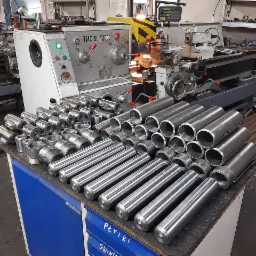 Stół warsztatowy z dużą ilością metalowych rur i elementów toczonych, w tle tokarka CNC TMC 50/2000 z włączonym panelem sterowania.