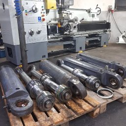 MAJKA Siłowniki - Cylindry hydrauliczne ułożone na palecie przed tokarką CNC w warsztacie, widoczne ślady użytkowania i drobne uszkodzenia na powierzchni.