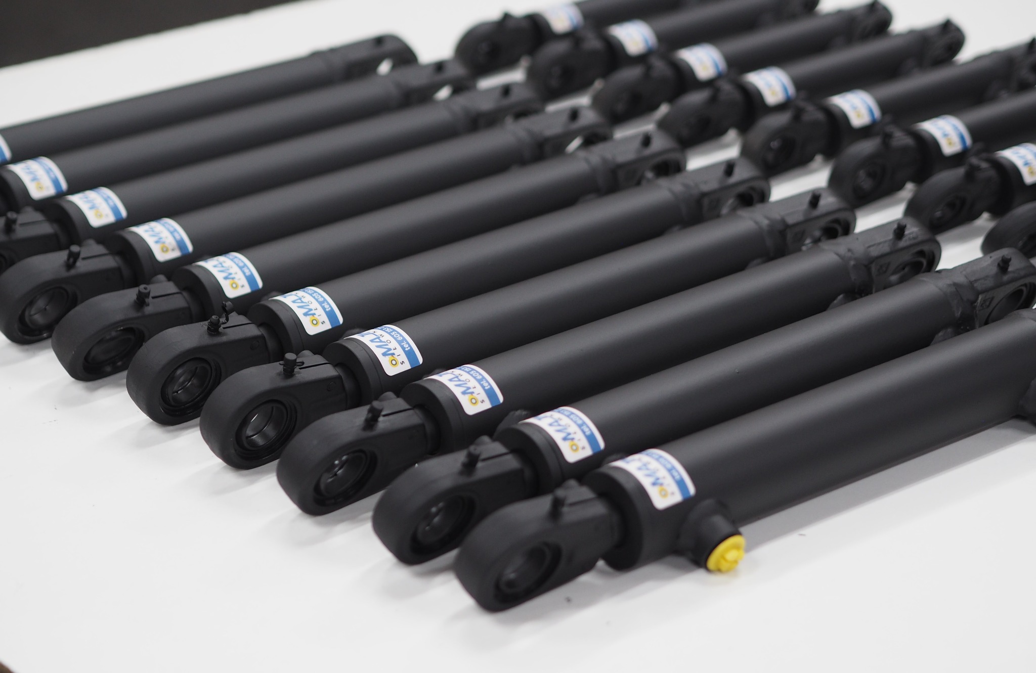Rząd cylindrów hydraulicznych w kolorze czarnym, z naklejkami producenta, ułożonych równolegle na białym tle, gotowych do montażu lub dystrybucji.