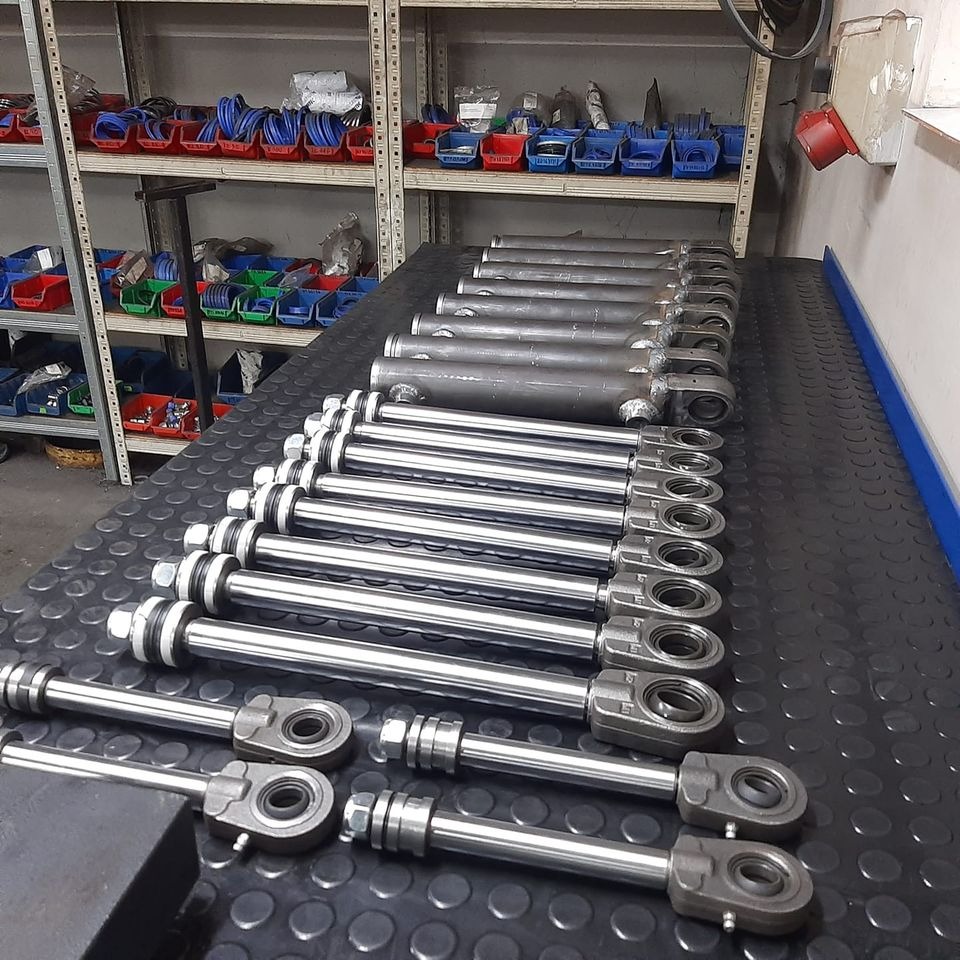 Rząd cylindrów hydraulicznych z polerowanymi tłoczyskami i szarymi uchwytami, ułożonych na czarnym blacie warsztatowym, z półkami wypełnionymi pojemnikami w tle.