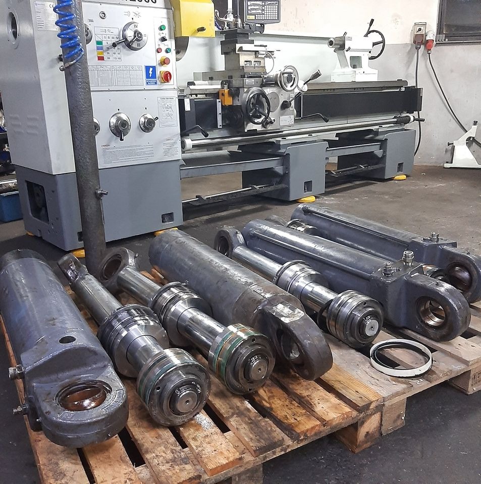 Cylindry hydrauliczne ułożone na palecie przed tokarką CNC w warsztacie, widoczne ślady użytkowania i drobne uszkodzenia na powierzchni.