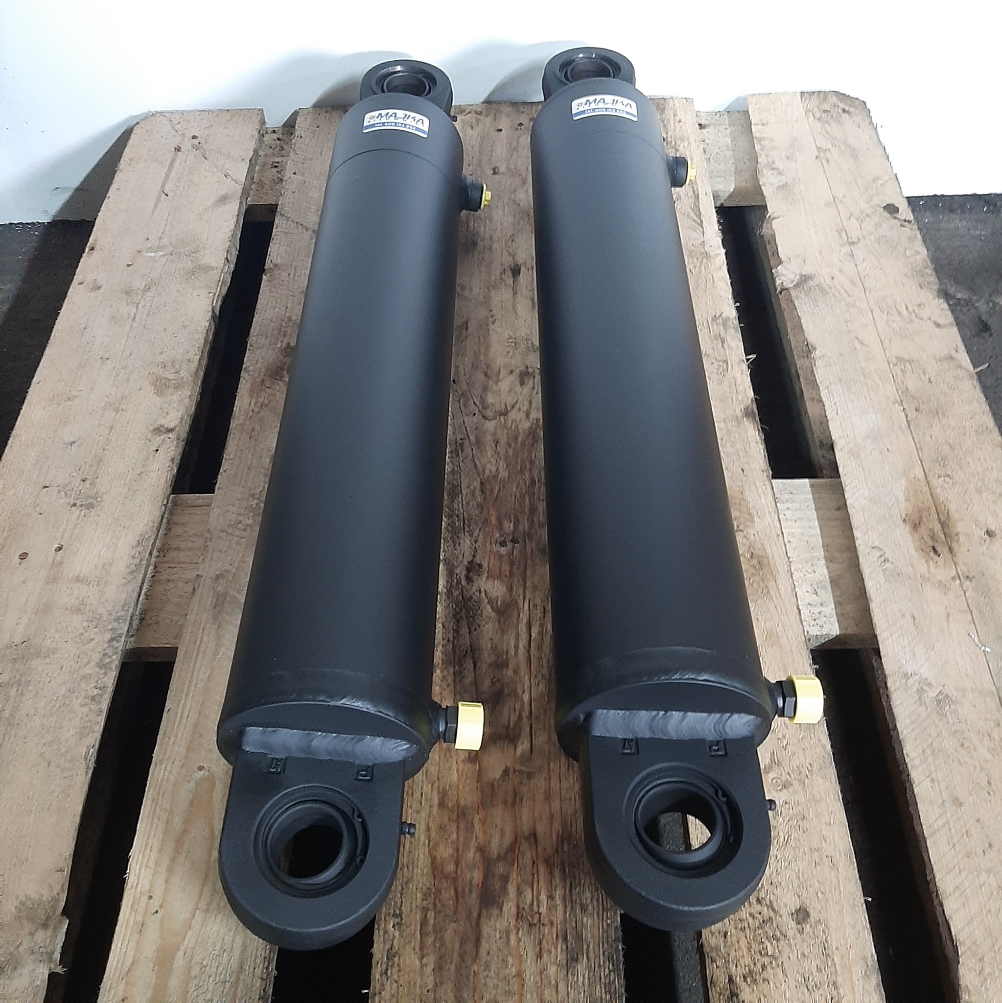 Dwa czarne cylindry hydrauliczne marki MAJKA, leżące na drewnianej palecie, widoczne z góry.