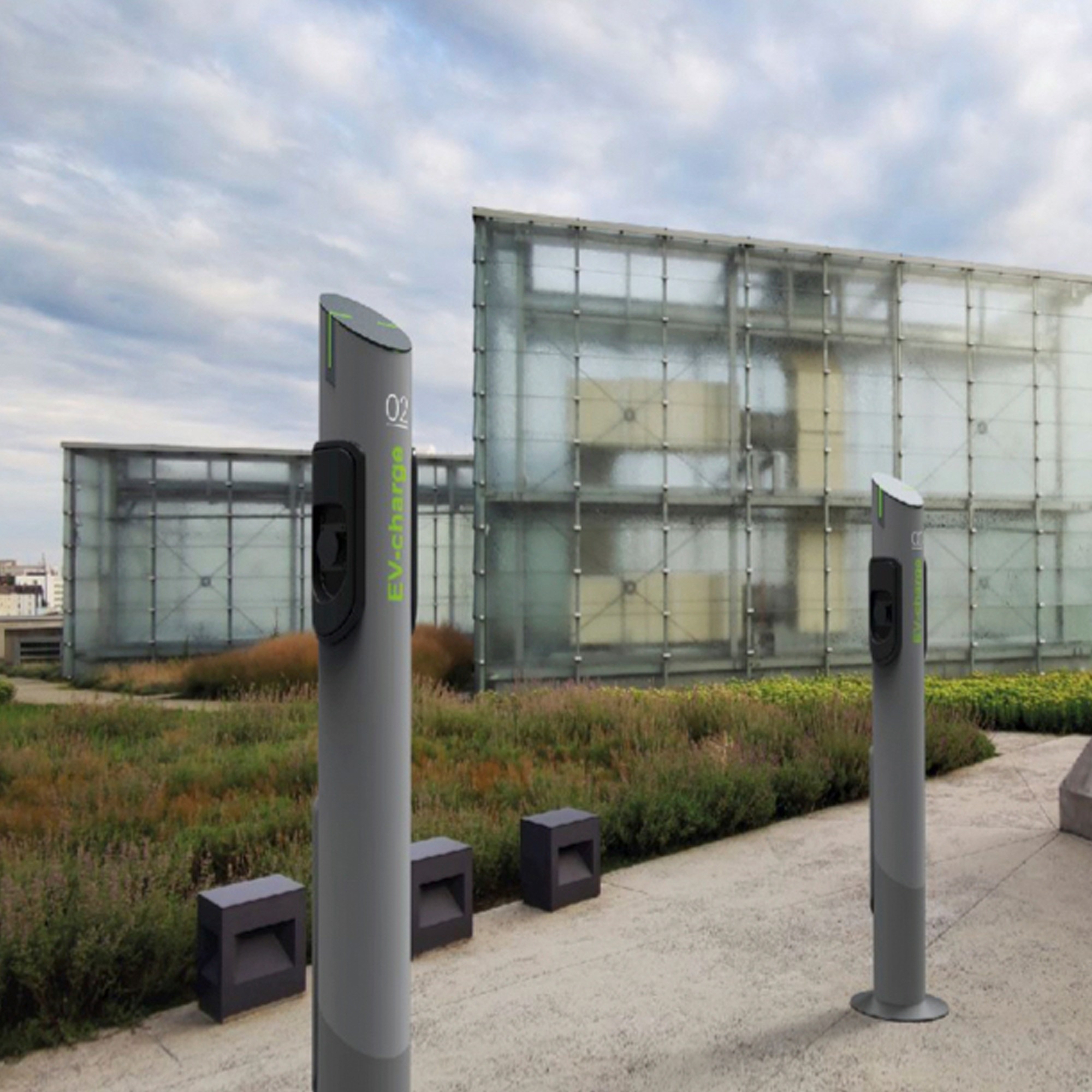 Dwie szare stacje ładowania pojazdów elektrycznych EV-charge na tle nowoczesnego budynku z przeszkloną fasadą i zielenią.