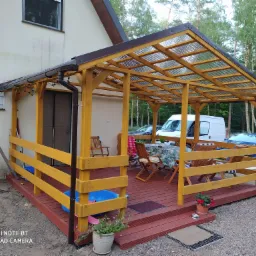 Żółta konstrukcja z przezroczystym zadaszeniem połączona z budynkiem, z drewnianym podestem, na którym stoją krzesła i stół nakryty obrusem, w tle las i zaparkowane samochody.